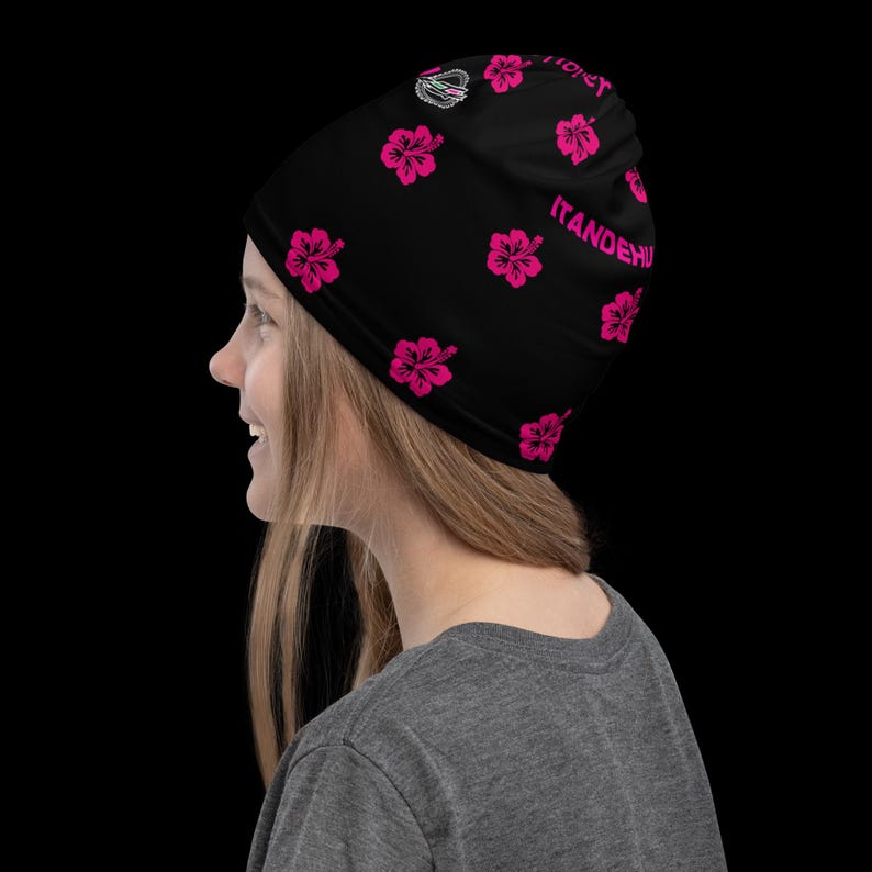May include: A black beanie featuring a repeating pattern of bright pink hibiscus flowers. The fabric displays the text ITANDEHUI in pink lettering along the side, with a small circular logo graphic positioned near the top crown of the headwear.