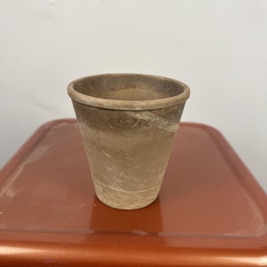 May include: A small, weathered terracotta pot with a slightly flared rim. The pot has a natural, earthy colour with variations in tone, from light beige to darker brown. The exterior shows subtle texture and signs of age, suggesting it's been used for planting.