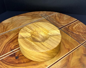 May include: A round wooden ashtray with a deep bowl, made from olive wood with a natural wood finish. The ashtray is sitting on a wooden surface with a decorative inlay.
