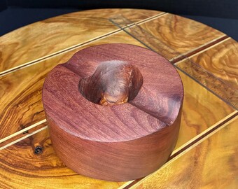 May include: A deep purple wooden ashtray with a rounded shape and a wide, shallow bowl. The ashtray is sitting on a wooden surface with a decorative inlay.