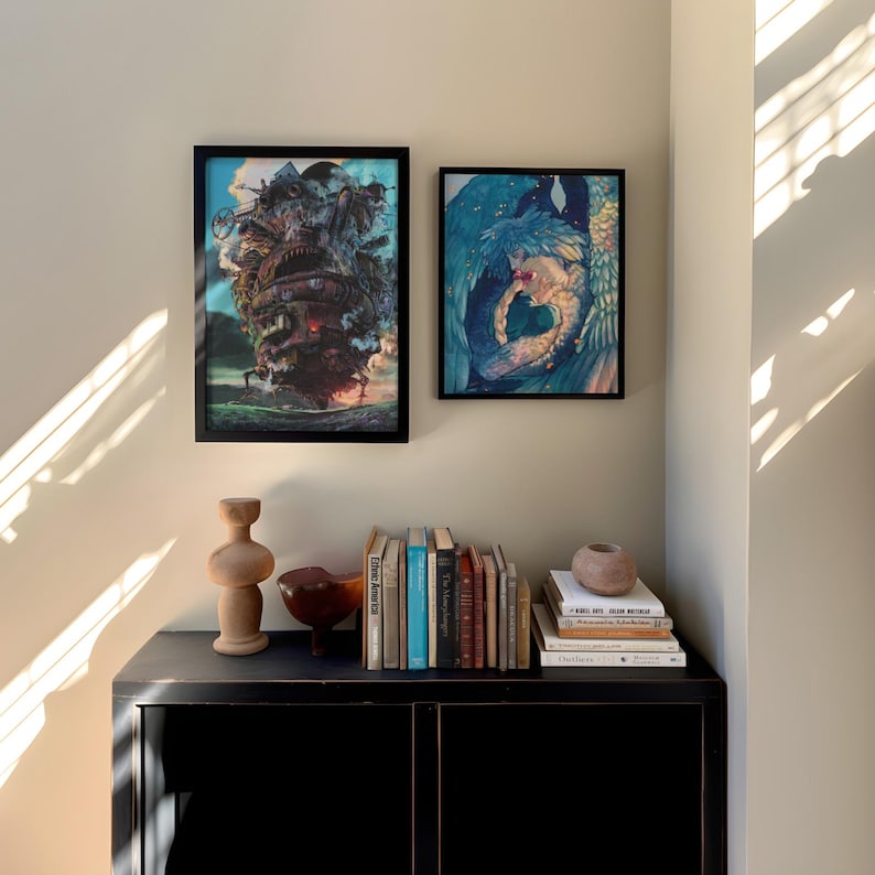 May include: Two framed art prints hang on a beige wall above a black cabinet. One print depicts a fantastical, detailed structure, while the other shows a figure with blue and white hues. A wooden sculpture, a bowl, and a stack of books are on the cabinet.