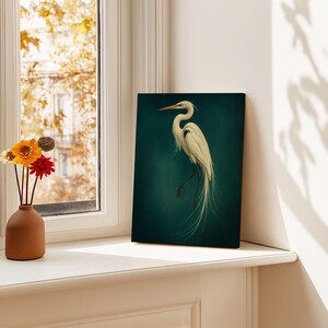 May include: A canvas print featuring a white egret against a dark teal background. The bird is depicted in a graceful pose, with long, flowing tail feathers. The artwork is displayed on a white windowsill, with a vase of flowers to the left.