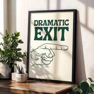 May include: A framed print featuring the words "DRAMATIC EXIT" in green, with a hand pointing right. The print has a cream background and a black frame. It's displayed on a wooden surface, alongside a potted plant and pencils in a holder.