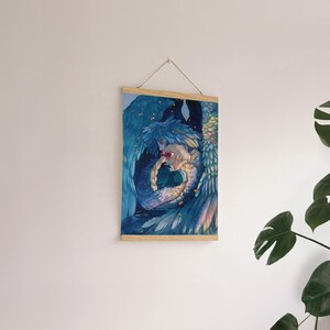 May include: A framed art print featuring a watercolor illustration of two figures embracing, surrounded by blue and teal wings. The artwork is mounted on a wooden frame and suspended by a string.