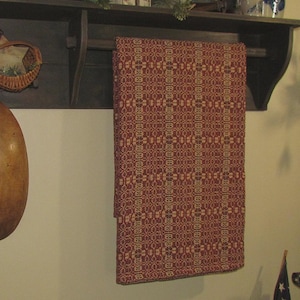 May include: A rectangular woven textile featuring a repeating geometric pattern in deep red and cream colors. The fabric is draped over a dark wooden rod attached to a wall-mounted shelf, displaying a consistent, intricate, and symmetrical design.