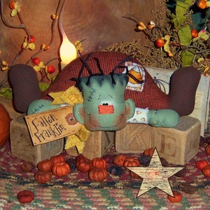 May include: A handmade Halloween decoration featuring a Frankenstein's monster doll with green skin, black hair, and stitched details. The doll is surrounded by miniature pumpkins, wooden blocks, and a star-shaped decoration. The sign reads "Fallen Franklin".