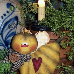 May include: A handmade felt bee with a black and white checkered scarf, a red heart, and a tag that says "Little Stitcher". The bee is sitting on a wooden surface with a lit candle and greenery in the background.