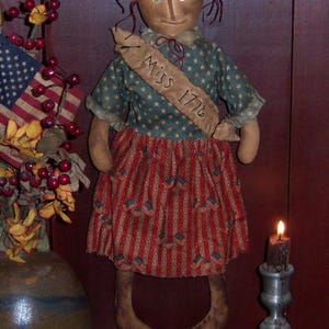 May include: A handmade rag doll dressed in a blue and white star patterned dress with a red and white striped skirt. The doll is wearing a sash that says "Miss 1776". The doll is standing in front of a wooden door with a sign that says "Pattern #591".