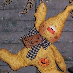 May include: A yellow fabric ghost with a black and white checkered bow and a tag that says "Scally" hanging from it. The ghost is decorated with the word "Boo" repeated many times. The ghost is hanging from a string and is in front of a wooden shutter.