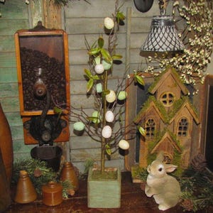 May include: A rustic Easter-themed display featuring a small tree adorned with speckled eggs in a square, light green planter. A decorative bunny figurine sits near a miniature house. A vintage coffee grinder and wooden accents complete the scene.