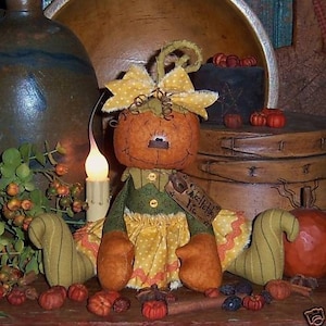 May include: A handmade pumpkin doll with a smiling face, wearing a green and yellow dress, and a yellow bow. The doll has orange arms and legs. The tag reads "Sweet Pumpkin Pie". Decorative pumpkins and a candle are in the background.
