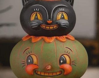 May include: A decorative figurine featuring a black cat head with a pink and orange party hat resting on a green pumpkin base. The pumpkin has a smiling face with orange cheeks and a nose, set upon an orange base with a diamond pattern and dots.