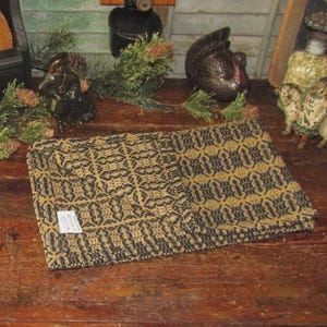 May include: A folded, rectangular textile with a repeating geometric pattern in black and tan. The fabric appears to be woven, with a label attached. The textile is displayed on a wooden surface, with decorative items in the background.