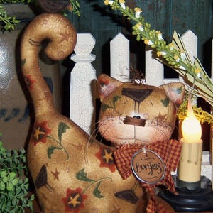 May include: A brown and tan cat doll with a floral pattern and a red and white checkered bow. The doll has a small bird perched on its tail and a tag that reads "Burgess".