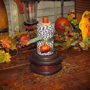 May include: A spool of black and white striped twine sits on a dark brown wooden base. A green ribbon is tied around the center of the twine, featuring a small, orange decorative pumpkin attached to the front of the spool as an accent piece.