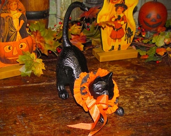 May include: A black cast iron cat figurine in a stretching pose, featuring a textured surface and a bright orange ruffled fabric collar with a matching satin ribbon bow tied around its neck, creating a decorative Halloween-themed accent piece.