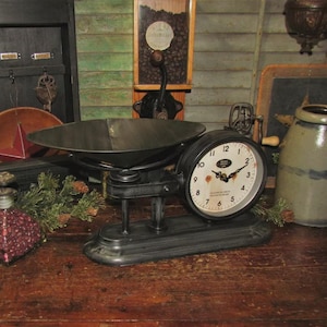 May include: Black vintage scale clock with a white face and black hands. The clock is set on a wooden surface with other antique items.