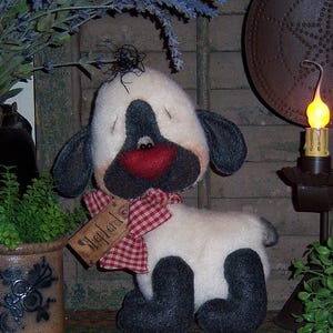 May include: A white and black sheep doll with a red and white checkered ribbon around its neck. The doll has a tag that says "Shepherd" on it. The doll is sitting on a brown surface.
