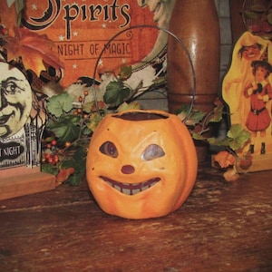 May include: A paper mache jack-o'-lantern with a wire handle. The jack-o'-lantern has a wide, toothy grin and large eyes. The jack-o'-lantern is orange and has a red nose. The jack-o'-lantern is sitting on a wooden table.
