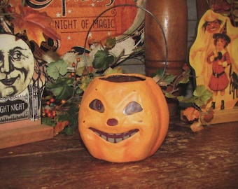 May include: A paper mache jack-o'-lantern with a wire handle. The jack-o'-lantern has a wide, toothy grin and large eyes. The jack-o'-lantern is orange and has a red nose. The jack-o'-lantern is sitting on a wooden table.