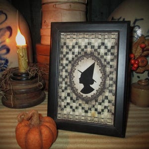 May include: A framed picture with a black frame featuring a witch silhouette. The background has a black and white checkered pattern. An orange pumpkin and a candle are in the foreground. The image has a rustic, vintage aesthetic.