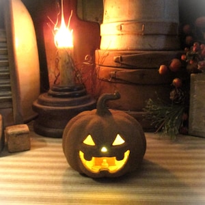 May include: A brown, textured jack-o'-lantern pumpkin figurine with a carved face featuring triangular eyes and a wide, toothy grin. The interior is illuminated with a warm, glowing yellow light, casting a soft radiance through the cutouts.