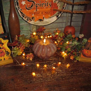 May include: A brown pumpkin-shaped candle with a lit flame, surrounded by a string of small glowing lights and artificial autumn leaves. To the left is a wooden cutout of a child in a witch costume, and to the right is a carved pumpkin cutout.