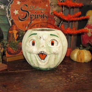 May include: A vintage, light green ceramic pumpkin-shaped container with a smiling face, orange eyes, and a jagged mouth. The container is part of a Halloween-themed display, with other decorative elements in the background.
