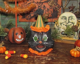 May include: A black cat-shaped Halloween candy bucket with a party hat and orange trim. The cat has green eyes and an open mouth. Other Halloween decorations include a pumpkin, candy corn, and a smiling moon.