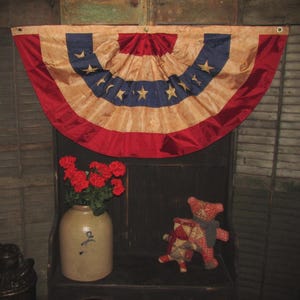 May include: A patriotic bunting in red, white, and blue, with gold stars, hangs above a dark wooden shelf. A ceramic jug with red flowers and a stuffed bear are on the shelf. The bunting is a decorative item.