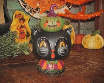 May include: A black cat figurine with large orange eyes and a surprised expression, wearing a striped orange and purple collar. A small green jack-o'-lantern with a purple stem sits balanced on top of the cat's head, completing the festive decor.