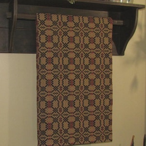 May include: A rectangular woven textile runner featuring a repeating geometric pattern in shades of tan, black, and deep red. The fabric displays a structured, grid-like design with intricate interlocking shapes across the entire surface area.