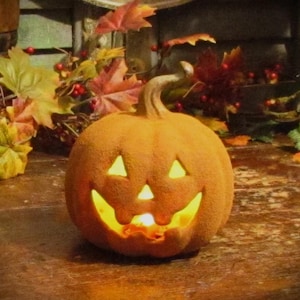 May include: A textured orange jack-o'-lantern pumpkin with a carved face featuring triangular eyes and a jagged mouth. The pumpkin is illuminated from within, casting a warm yellow glow through the cutouts, creating a festive autumn display.