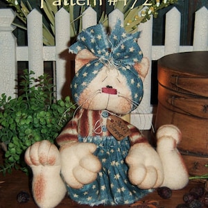 May include: A handmade rag doll cat wearing a blue and white star patterned dress and a matching headband. The doll has a red nose and white fur. The doll is sitting on a wooden surface with cinnamon sticks and berries.