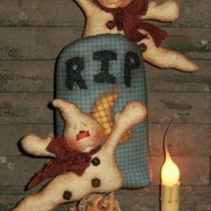 May include: Halloween-themed fabric figures with a tombstone that reads "RIP". The figures are cream-colored with red bows and button accents. A lit candle and small pumpkins complete the spooky scene, perfect for Halloween decor.