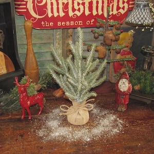 May include: A small, artificial Christmas tree with frosted green needles sits in a burlap sack tied with twine. A red reindeer figurine and a carved Santa Claus figure are on a wooden surface. A sign that says "Christmas" is in the background.