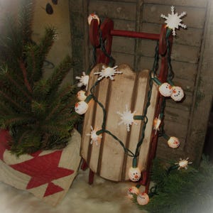 May include: A rustic wooden sled with red runners is adorned with a string of snowman and snowflake lights. A small Christmas tree sits to the left in a red star-patterned fabric pot. The scene evokes a cozy winter holiday theme.