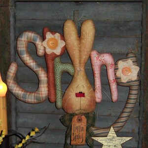 May include: A handmade spring-themed decoration with the word "Spring" spelled out in fabric letters. The letters are decorated with plaid, floral, and striped patterns. A bunny with a red nose is in the center of the word. The decoration is hanging from a wooden stick with a green and brown plaid ribbon and a tag that says "Hoppin' Spring". A gold star is on the bottom right of the image.