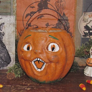 May include: A vintage orange pumpkin-shaped Halloween candy pail with a wire handle. The pumpkin has a carved face with white eyes, a toothy grin, and green accents. Other Halloween decorations are in the background, including a witch and a ghost.