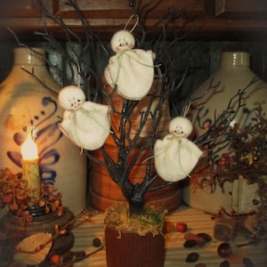 May include: A Halloween-themed centerpiece featuring a small, dark-colored tree adorned with three white ghost ornaments. The tree is potted in a brown container and surrounded by decorative elements like cinnamon sticks and dried leaves. The background includes antique jugs and a candle.