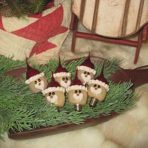 May include: A collection of six Christmas light bulbs decorated as Santa Claus figures. Each bulb has a red hat with glitter, a white beard, and a smiling face. They are arranged on a wooden scoop with green pine needles.