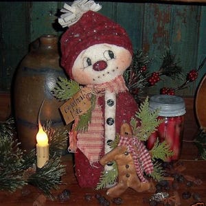 May include: A handmade snowman doll with a red hat, white face, and a red and white striped scarf. The snowman is holding a gingerbread man cookie. The doll is sitting on a wooden shelf with a jar of red berries and a sign that says "Gingerbread Lane".