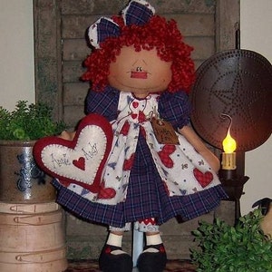 May include: A handmade doll with red yarn hair, wearing a plaid and heart-patterned dress. The doll holds a heart-shaped pillow with the words "Annie Andy" on it. The doll has black shoes and a matching bow.
