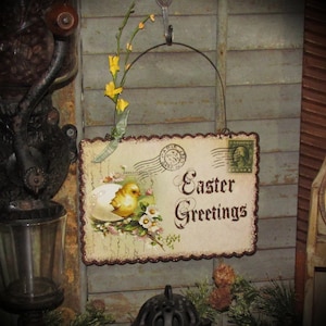 May include: A vintage-style Easter sign with the words "Easter Greetings" in elegant script. The sign features a charming illustration of a baby chick hatching from an egg, surrounded by flowers. The sign is decorated with a scalloped edge and a decorative border.