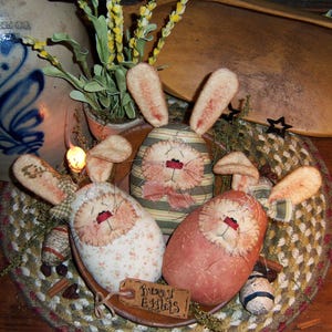 May include: Four handmade bunny-shaped Easter eggs in a woven basket. The eggs are made of fabric and have embroidered faces. The eggs are decorated with different patterns and colors, including pink, green, and white. The basket is surrounded by dried flowers and other Easter decorations.