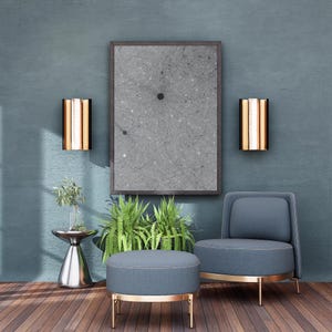 May include: A modern interior scene featuring a gray abstract art print in a dark wood frame, flanked by two gold and copper wall sconces. A blue armchair and ottoman with gold trim sit on a wooden floor, with plants adding a touch of nature.