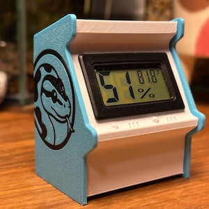 May include: A small, retro-style digital display unit with a light blue and white casing. The screen shows 51% humidity and 81.8°F. A black and white snake logo is on the side.