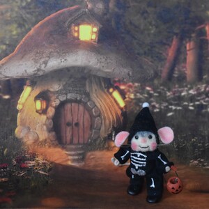 May include: A small, gray mouse wearing a black skeleton costume and a black hat is holding a small orange pumpkin bucket. The mouse is standing in front of a miniature mushroom house with a lit window and a wooden door. The house is surrounded by green foliage and flowers.