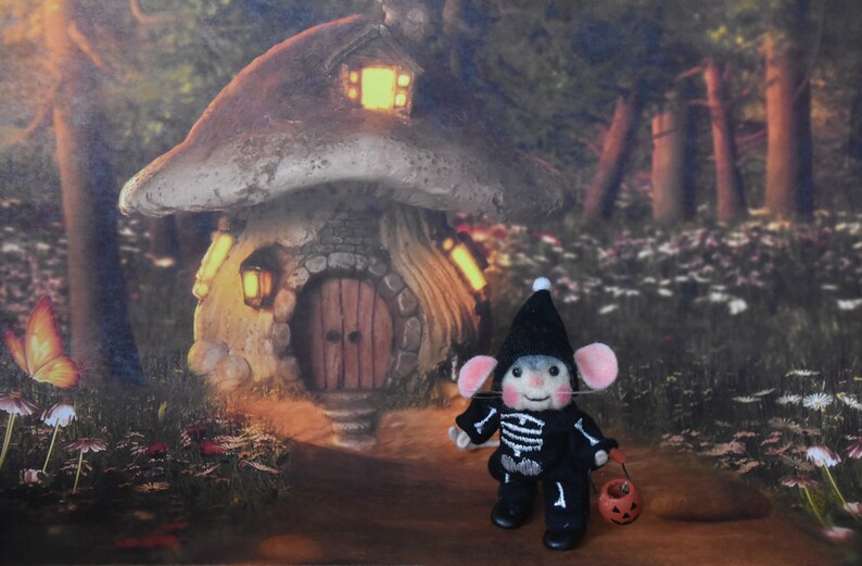 May include: A small, gray mouse wearing a black skeleton costume and a black hat is holding a small orange pumpkin bucket. The mouse is standing in front of a miniature mushroom house with a lit window and a wooden door. The house is surrounded by green foliage and flowers.