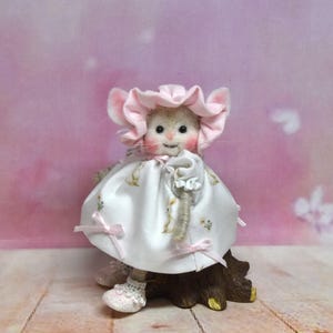 May include: A handmade mouse figure, dressed in a pink bonnet and a white dress adorned with a floral design. The mouse is positioned on a brown base, with pink bows on the dress and shoes. The backdrop is a soft pink hue.
